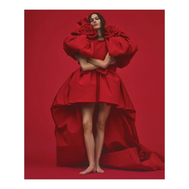 A woman in a red couture gown with large roses at the shoulders stands in a photo studio with a red background for a fashion shoot.
