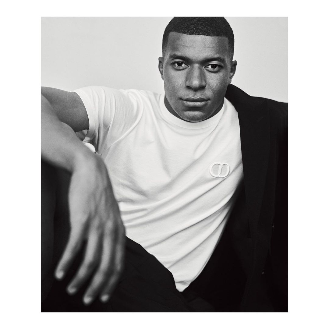 Kylian Mbappé wearing a white t-shirt and dark jacket against a plain background in a portrait photograph by Gregory Harris.