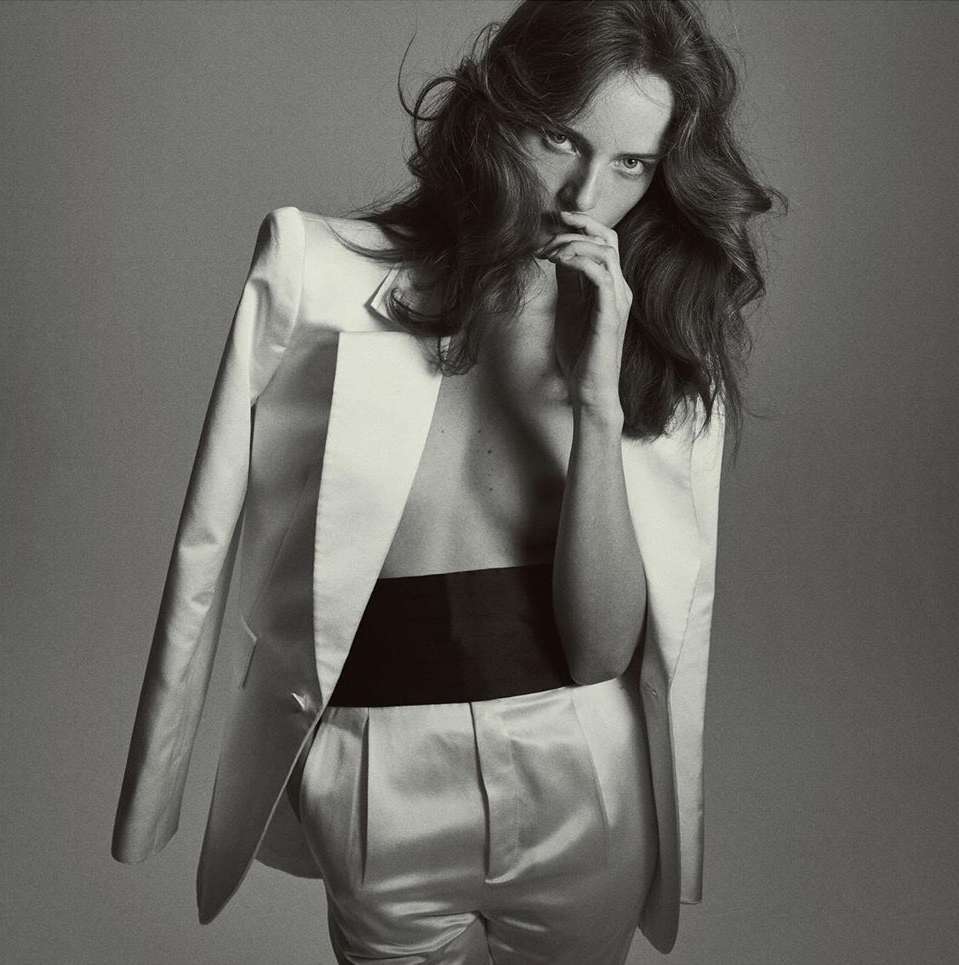 Monochrome portrait of a woman with long, wavy hair wearing a white blazer and pants with a black waistband, striking a pose indoors.