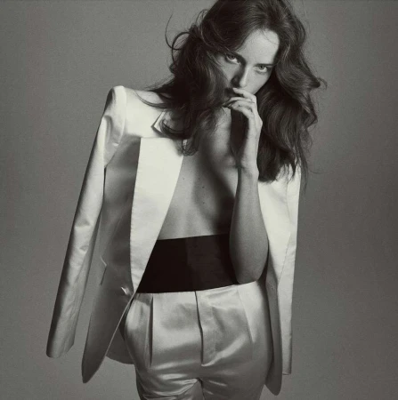 Monochrome portrait of a woman with long, wavy hair wearing a white blazer and pants with a black waistband, striking a pose indoors.