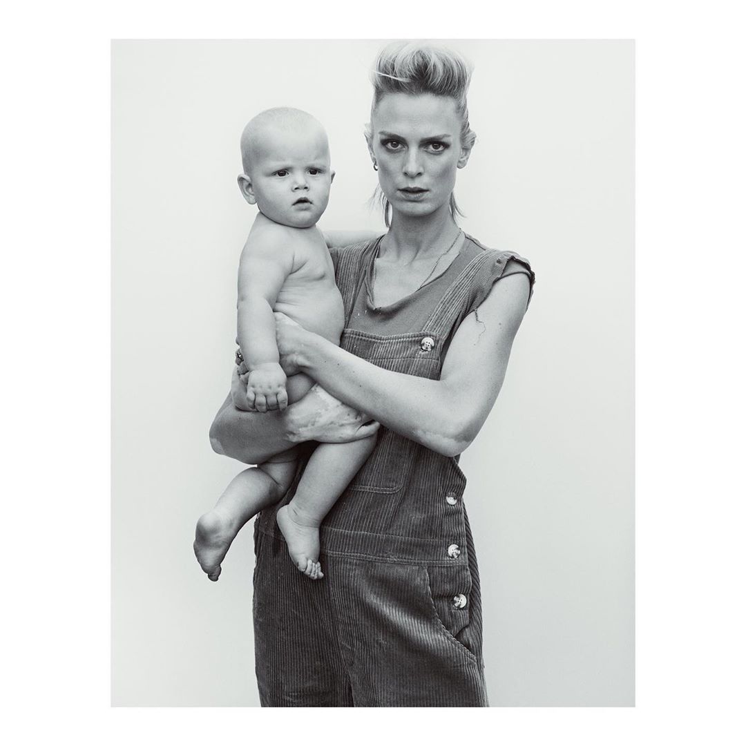 Woman in overalls holding a baby against a plain light background photographed in New York by Gregory Harris