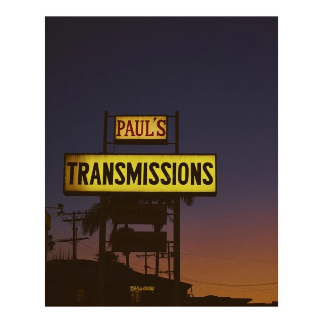 Illuminated sign reading "Paul's Transmissions" at dusk, with a gradient sky, street elements, and silhouettes of buildings and palm trees.