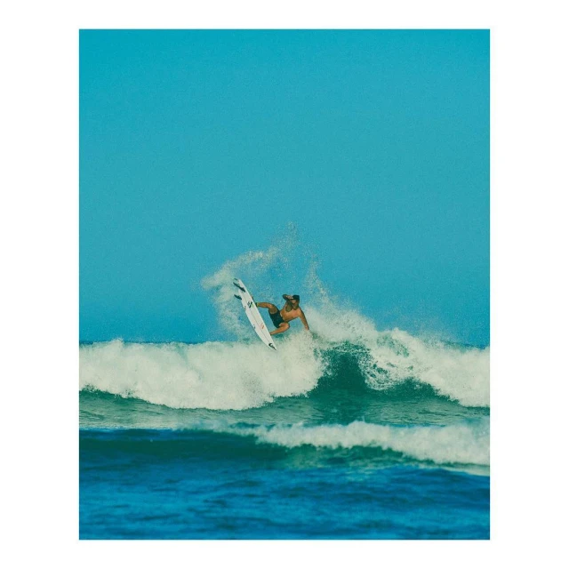 Male surfer riding a wave on a surfboard in an ocean environment under a clear blue sky with splashing white water.