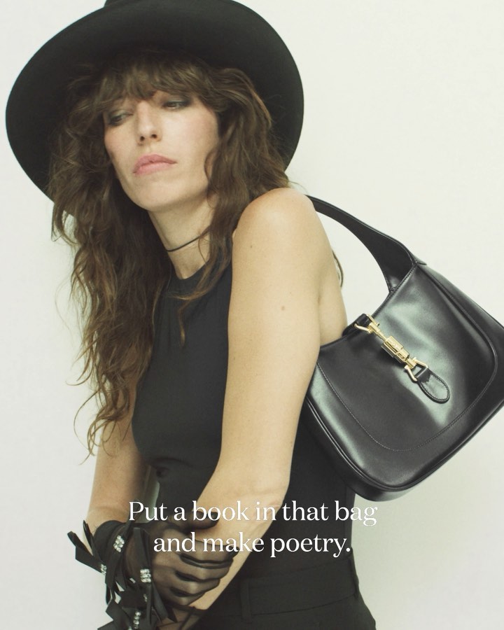A woman in a black hat poses with a black leather purse in a studio setting, showcasing fashion and style.