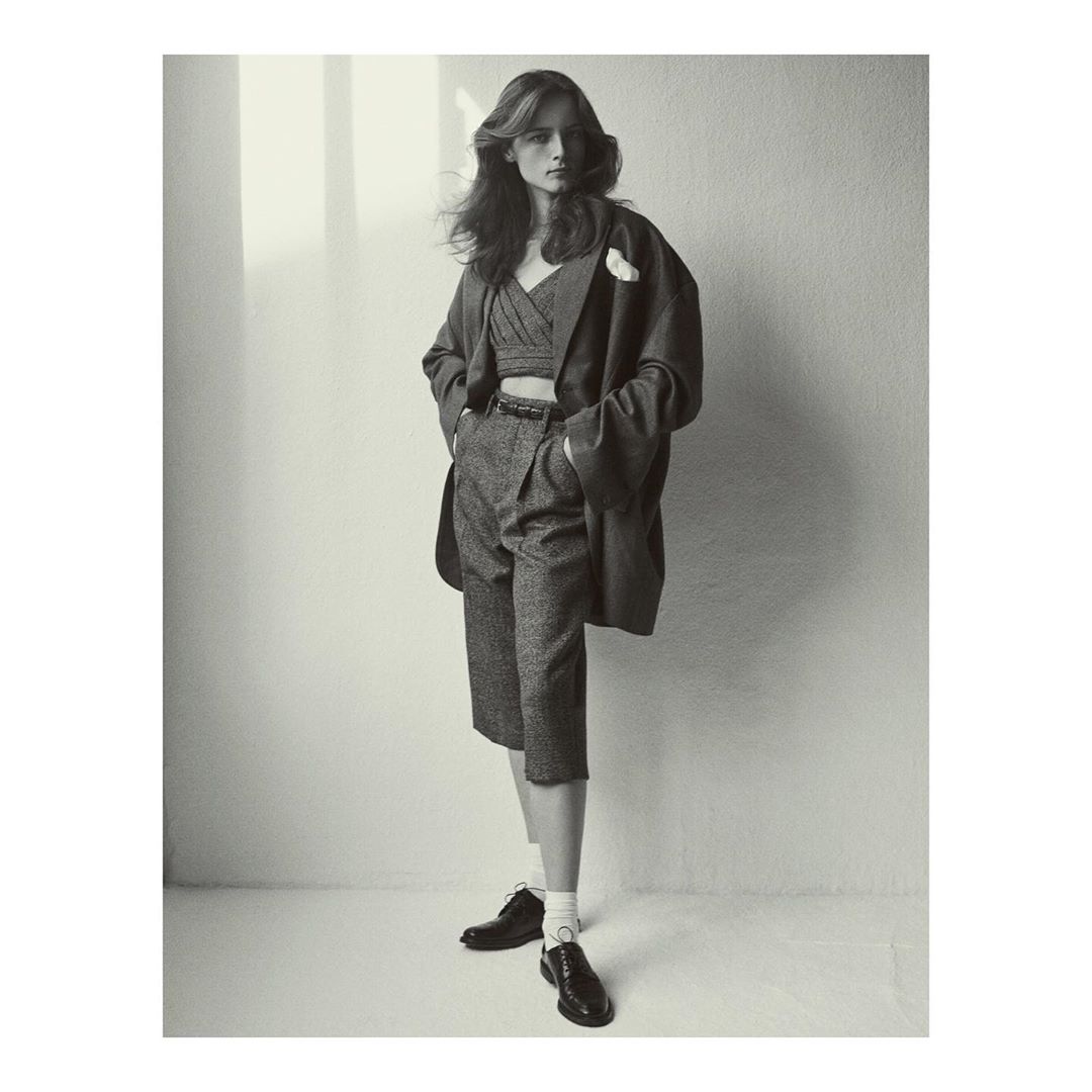 A woman poses in a cropped top, culottes, a blazer, and dress shoes in a studio with natural light and shadows on the backdrop.
