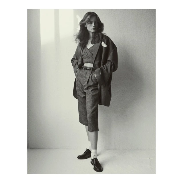 A woman poses in a cropped top, culottes, a blazer, and dress shoes in a studio with natural light and shadows on the backdrop.