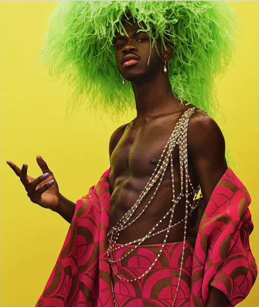 Adult male with green feathered headpiece in front of yellow backdrop wearing beaded necklaces and pink patterned robe.