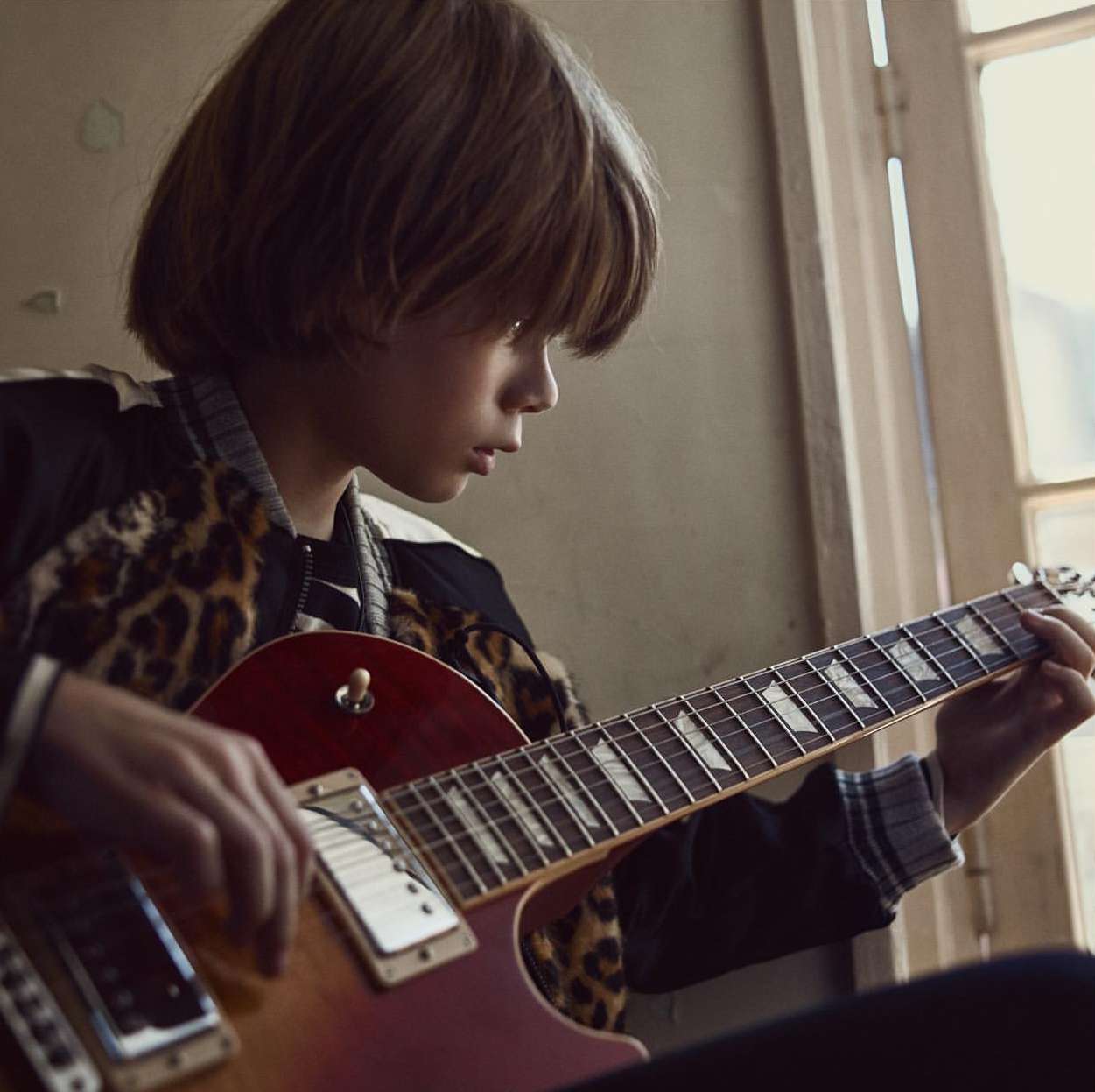 A young boy with brown hair plays an electric guitar indoors by the window, wearing a leopard-print jacket. Medium shot, eye-level angle.