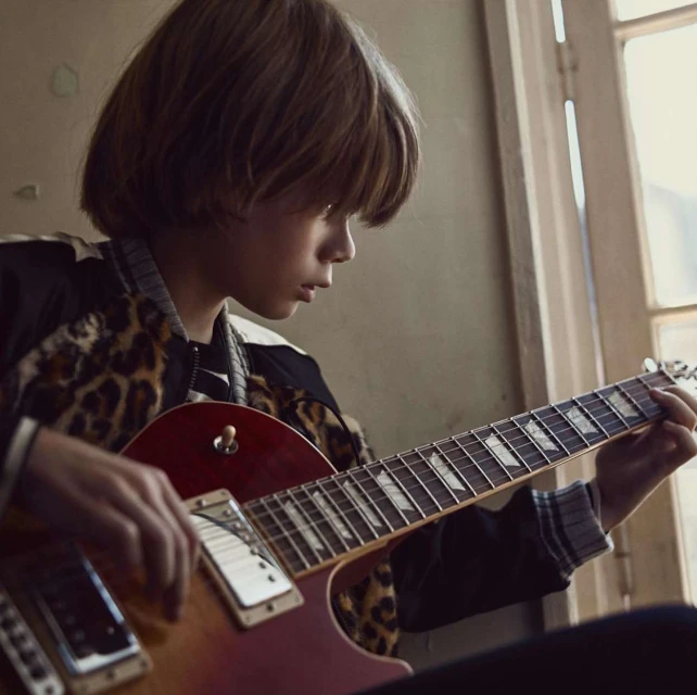 A young boy with brown hair plays an electric guitar indoors by the window, wearing a leopard-print jacket. Medium shot, eye-level angle.