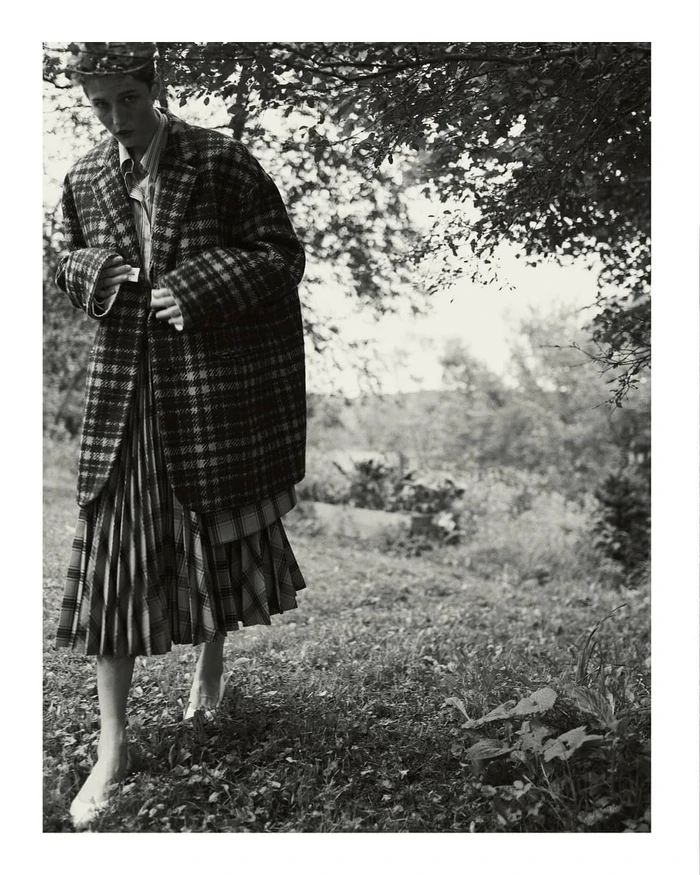 Model in plaid oversized jacket and pleated skirt walking outdoors through a grassy field under tree branches.