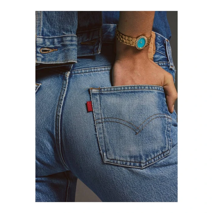 Close-up of a person wearing denim jeans and jacket, with a gold watch featuring a turquoise dial on the wrist.