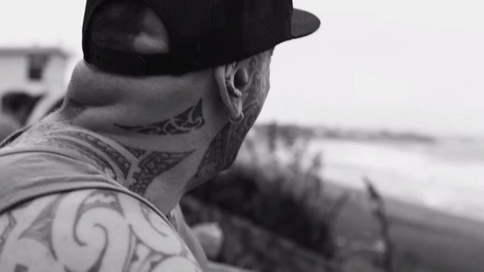 Man wearing a baseball cap with extensive neck and arm tattoos looking out toward the ocean