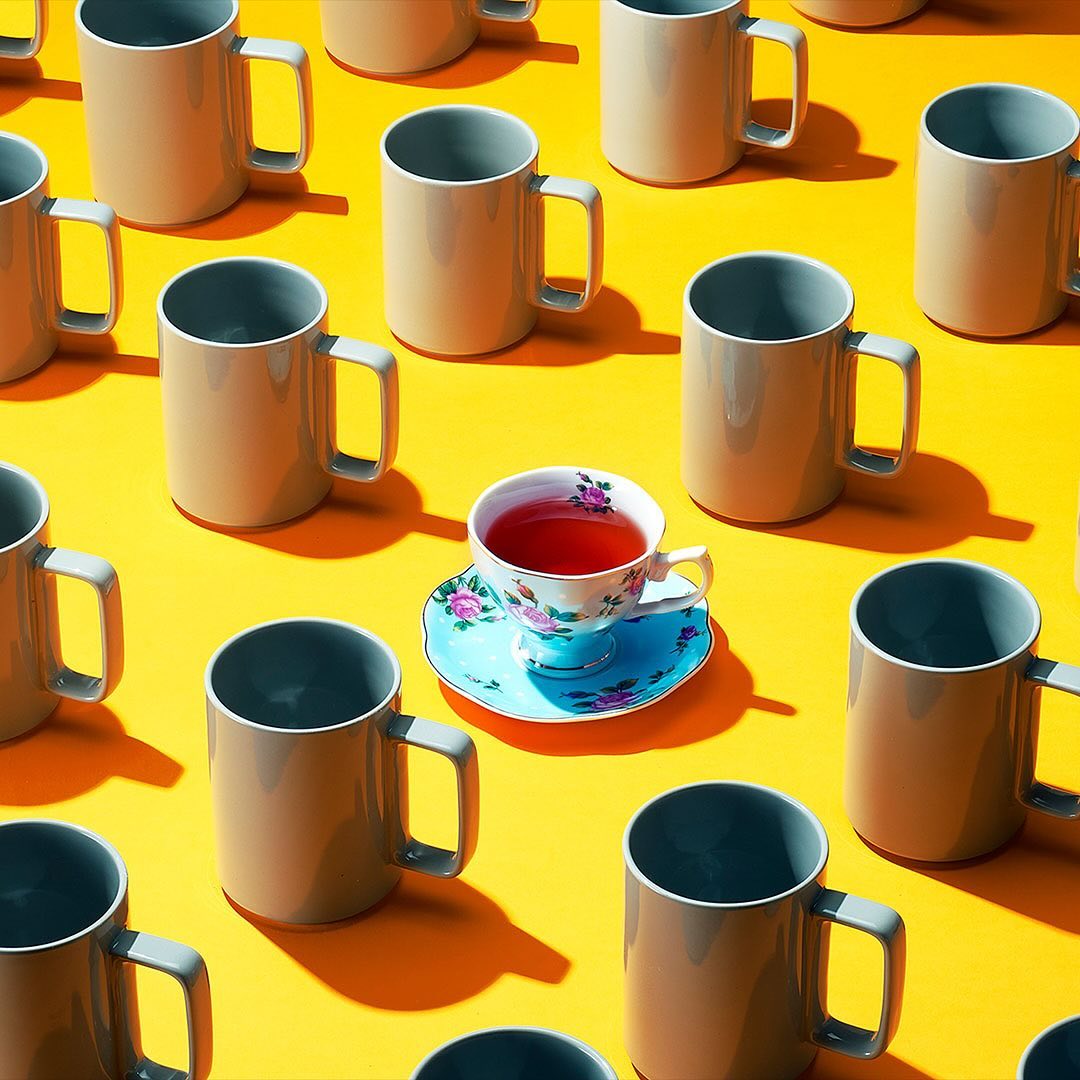 Elevated, grid-like pattern of beige coffee mugs and an ornate teacup filled with tea against a vivid yellow background.