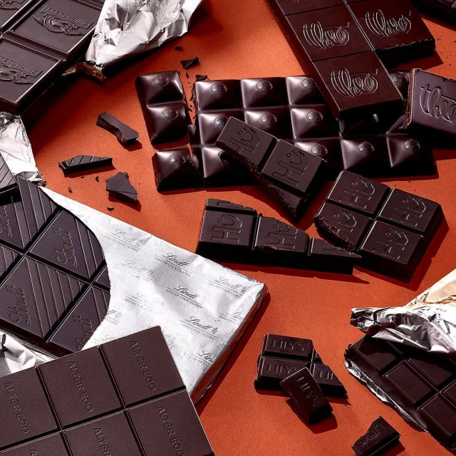 Still life shot of dark chocolate bars and pieces on a reddish-brown surface with silver foil wrappers, creating a sweet and decadent scene.