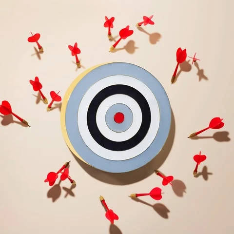 Overhead shot of a dartboard with concentric circles surrounded by red darts, some hitting the board and others scattered around it.
