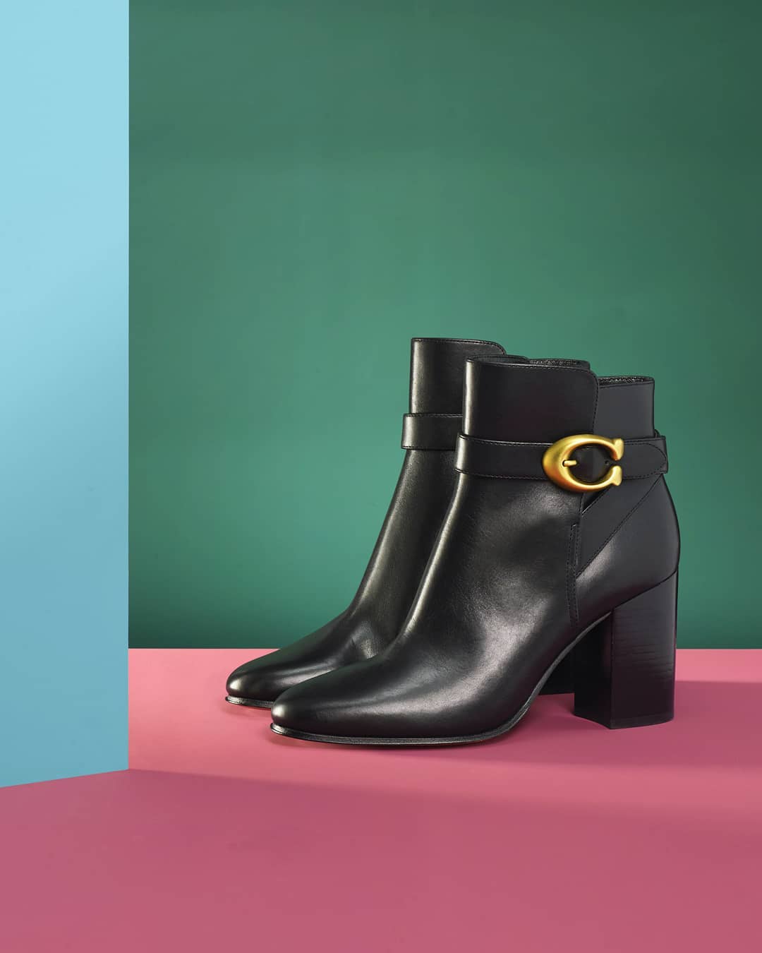 Pair of black leather ankle boots with gold buckle detail. Fashion footwear photography against green and pink backdrop.