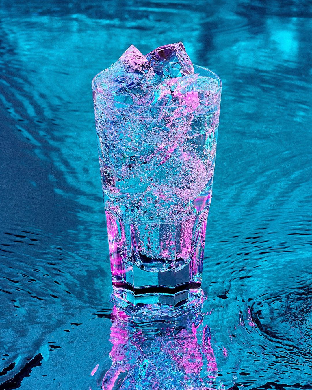 Tall glass filled with ice and a clear liquid sits partially submerged in blue water creating vibrant reflections and refractions.