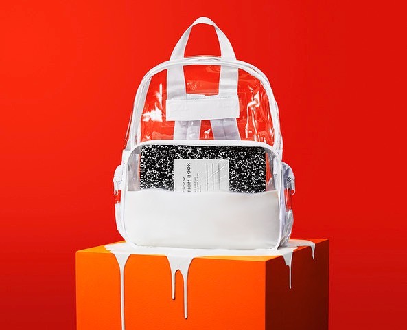 Clear backpack on an orange cube against a red backdrop in a studio shot featuring white paint dripping down the sides.