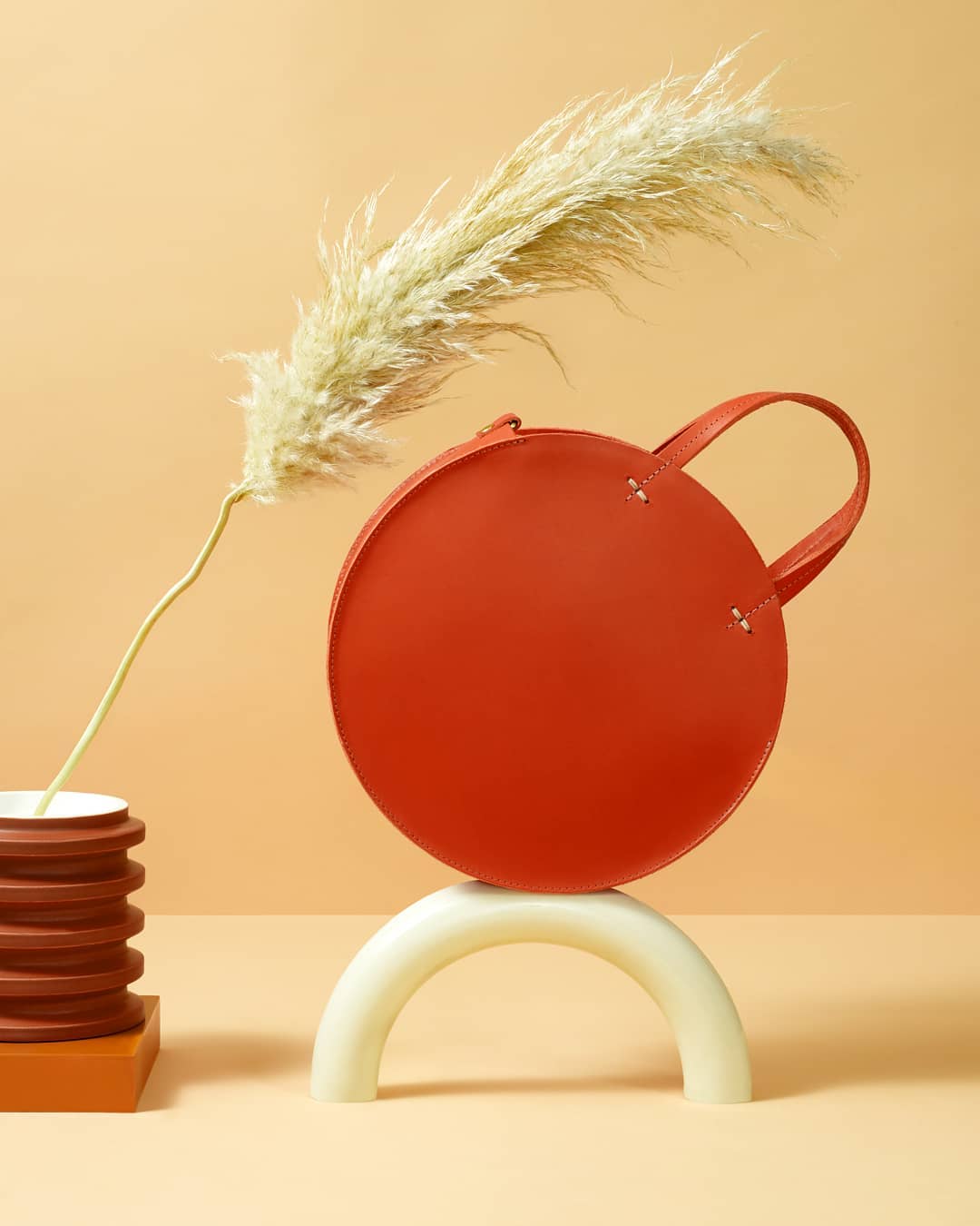 Studio shot of an orange circular handbag balancing on a white arc with pampas grass and brown ribbed vase against peach background.