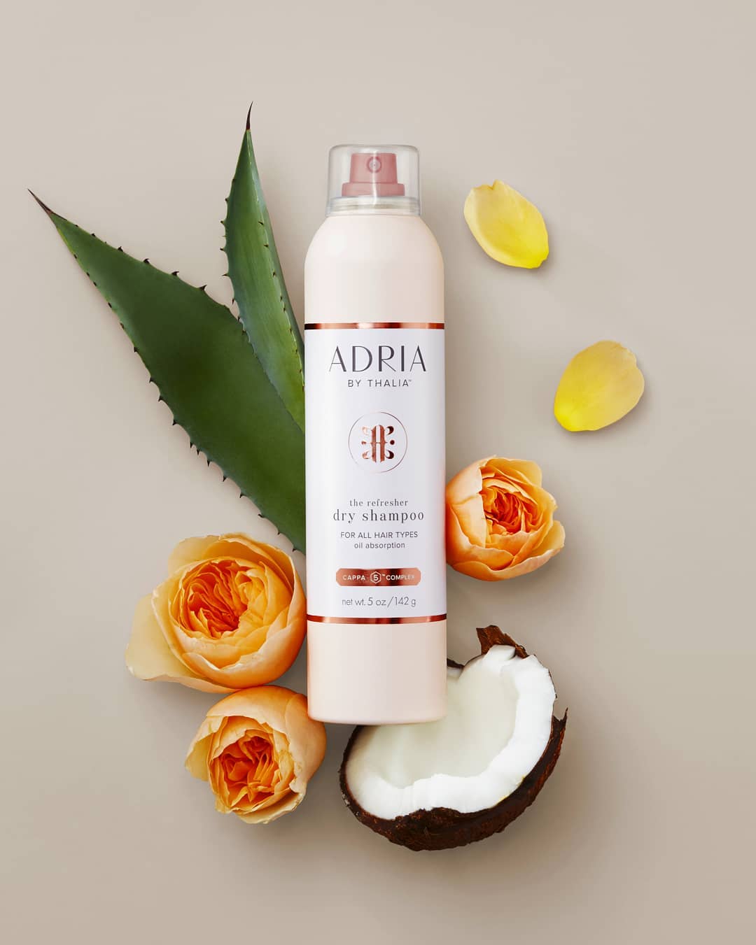 Dry shampoo by Adria, with aloe vera leaves, roses and coconut arranged against a beige background in a beauty product still life.