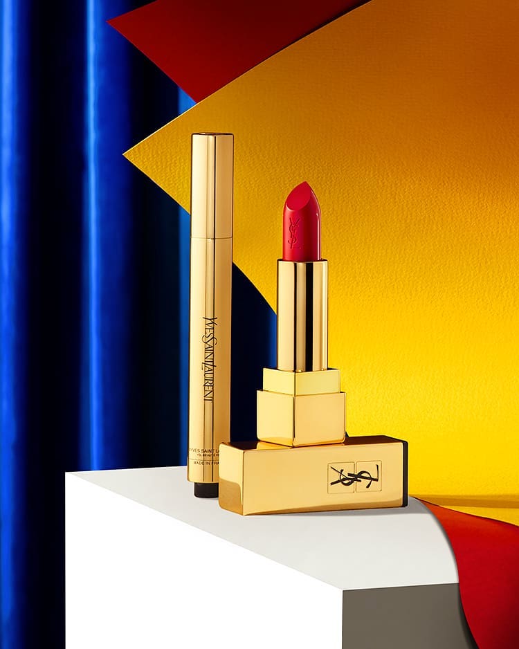 Red lipstick and golden cosmetic tube are displayed on a white platform against a vibrant background of blue, yellow, and red.