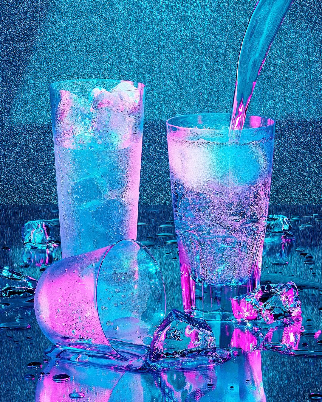Icy drink photography: liquid being poured into a tall glass filled with ice next to two other glasses on a wet surface.