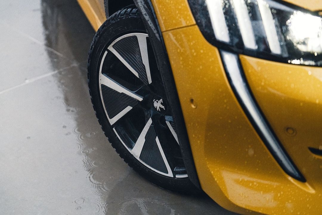 Close-up shot of a yellow Peugeot car's front wheel, tire, and headlight on a wet surface, highlighting automotive design details.