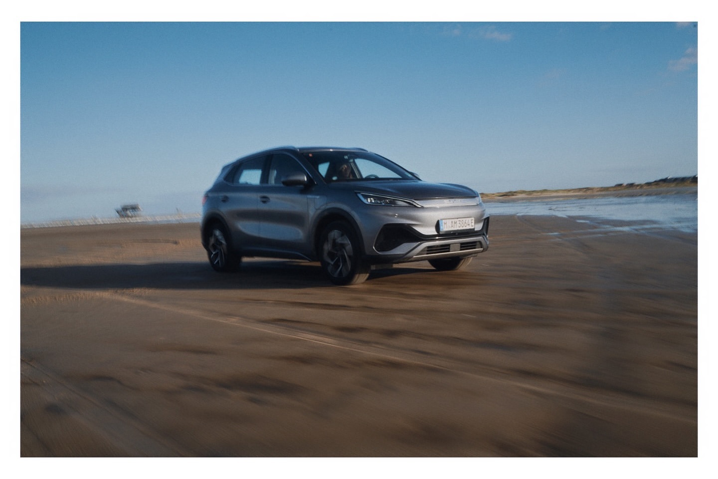 Silver BYD Atto 3 electric SUV driving on a sandy beach with a clear blue sky, showcasing automotive design and a coastal environment.