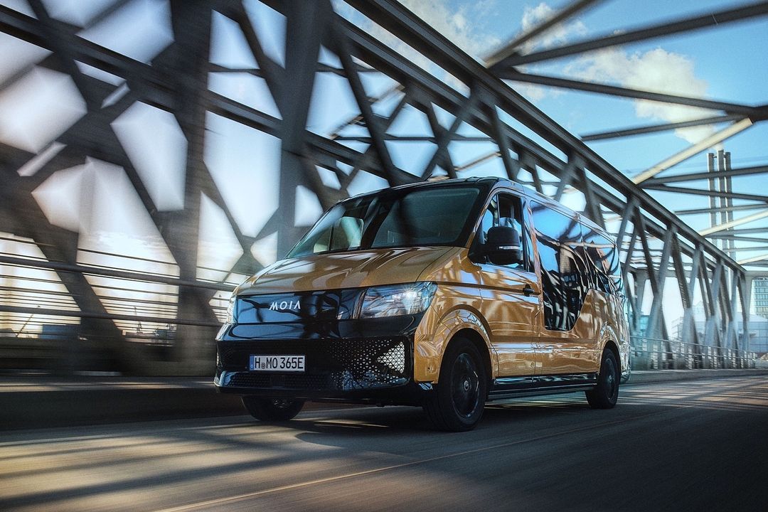 A golden MOIA passenger van drives across a bridge on a sunny day, showcasing its modern design and urban mobility.