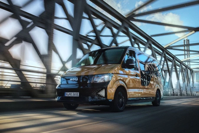 A golden MOIA passenger van drives across a bridge on a sunny day, showcasing its modern design and urban mobility.