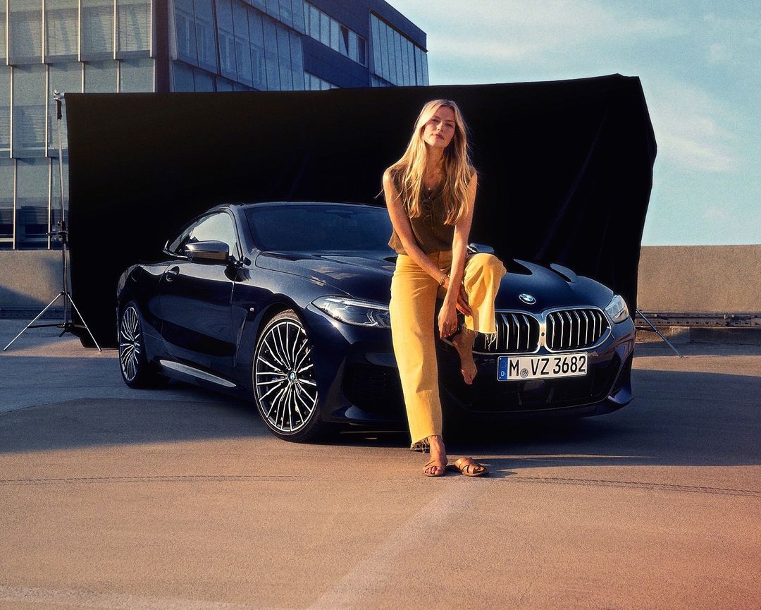 Woman in yellow pants sits on the hood of a dark blue BMW in an outdoor setting with a photo backdrop, automotive scene.