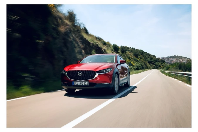 Red Mazda CX-30 driving on a sunny road surrounded by green hills and trees under a bright blue sky, capturing a sense of motion.