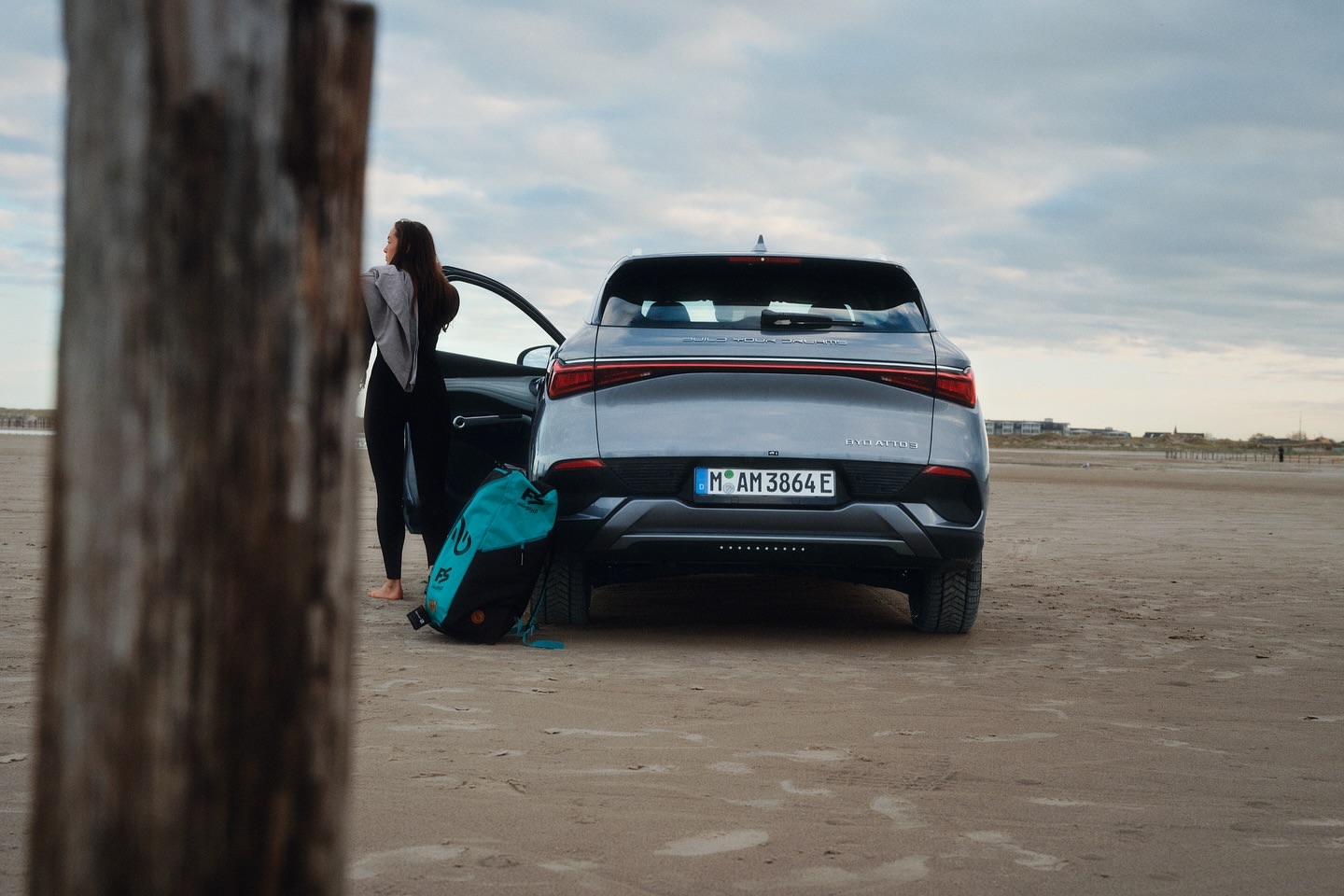 Rear of a gray electric SUV on a sandy beach near the ocean, parked with its door open and a person and surfboard bag nearby.