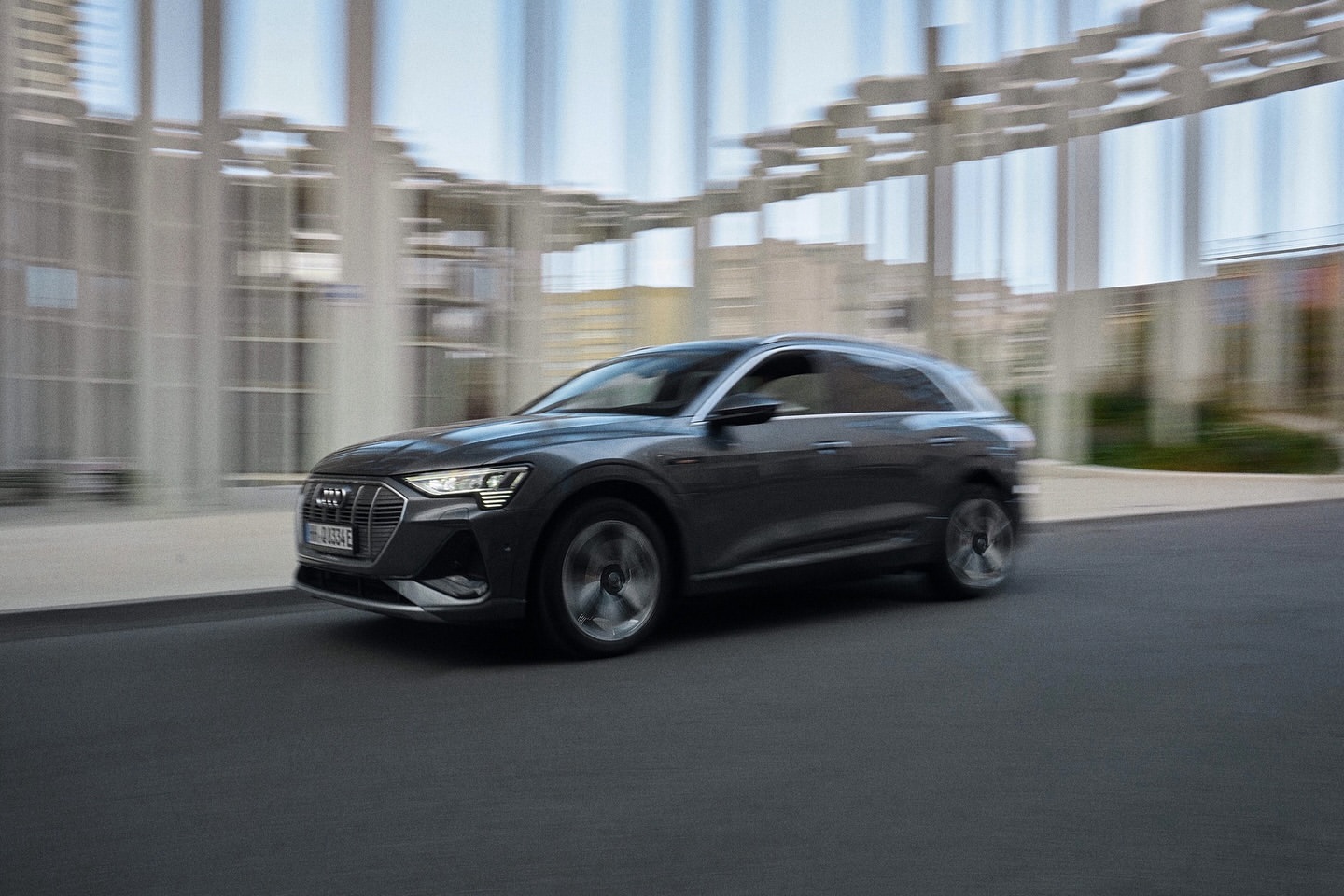 Grey Audi e-tron driving on paved road in an urban environment; car is in motion with building in background.