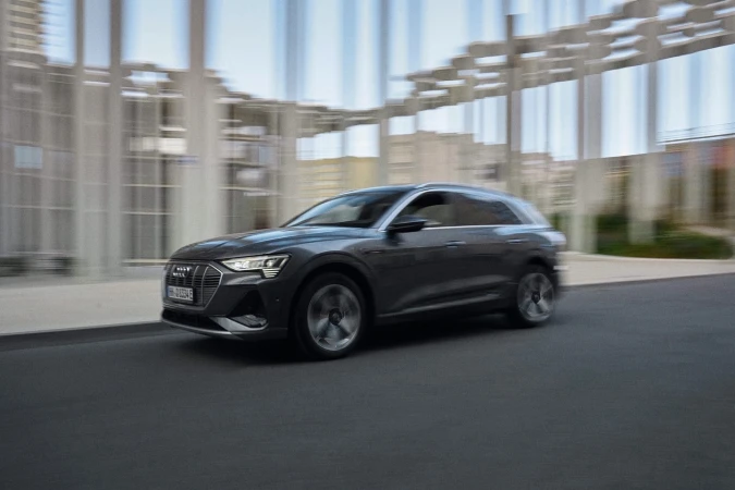 Grey Audi e-tron driving on paved road in an urban environment; car is in motion with building in background.