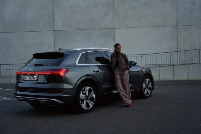 Woman in tailored suit leans against grey Audi e-tron electric SUV parked outdoors against a concrete wall backdrop. Automotive fashion.