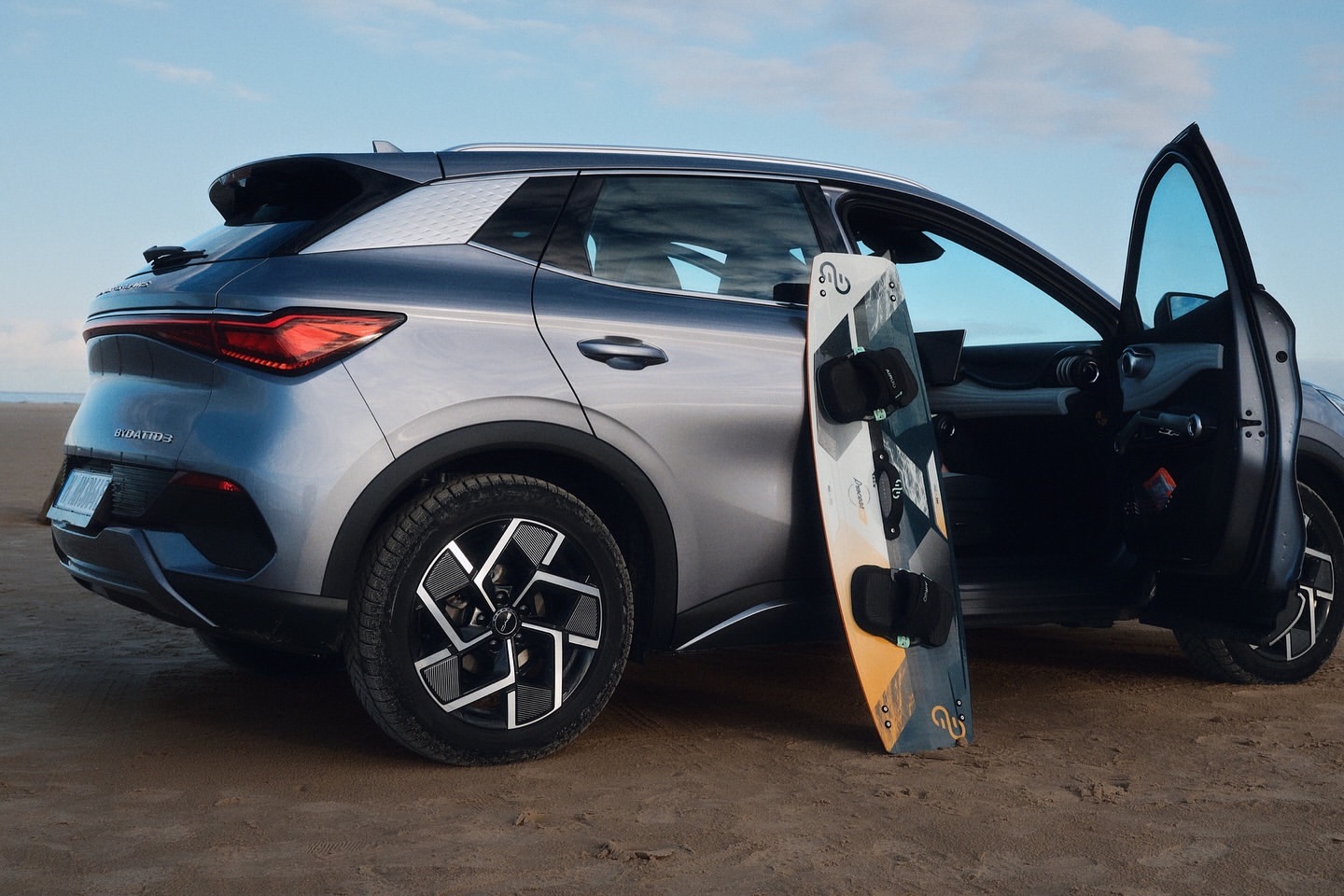 Silver SUV parked on sandy beach with open door and wakeboard, under a blue sky. Automotive, travel, and outdoor adventure.