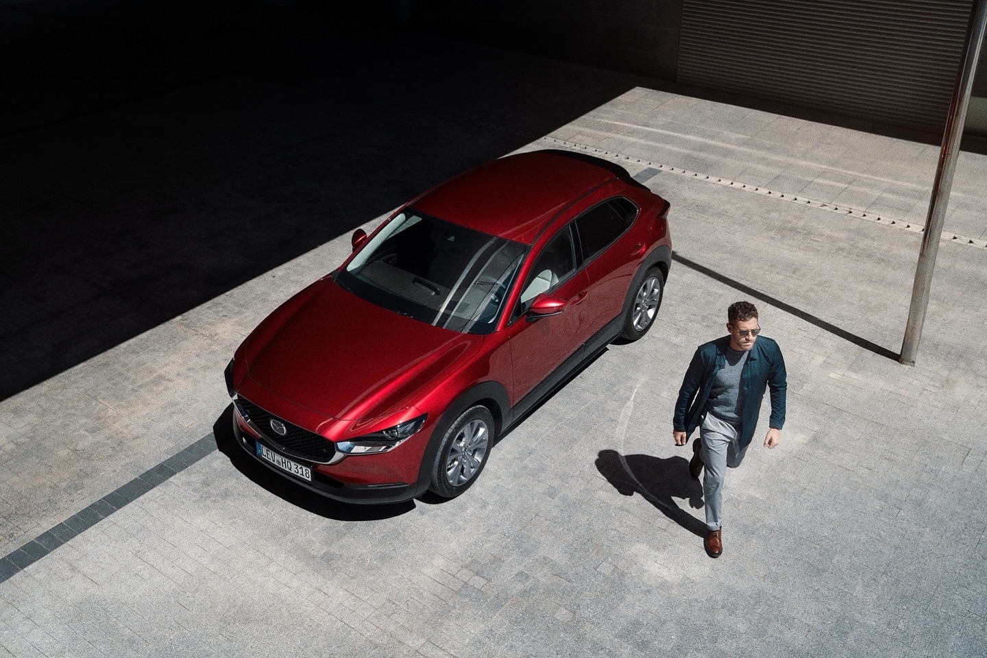 Overhead view of a red Mazda CX-30 parked on concrete, with an adult male walking nearby in daytime, creating shadows.