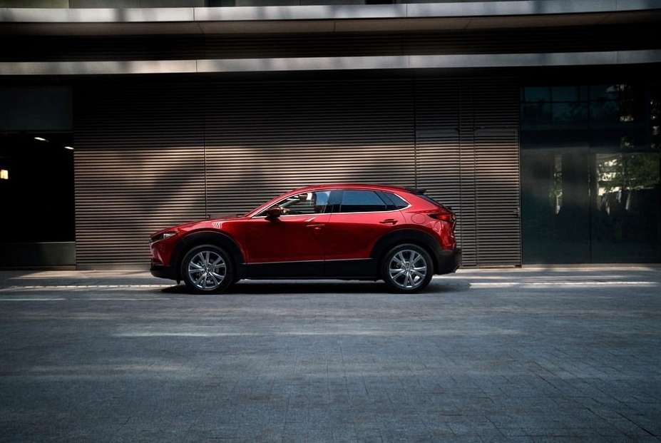 Red Mazda CX-30 parked on asphalt street against a modern building with horizontal lines and glass doors, captured in an eye-level shot.
