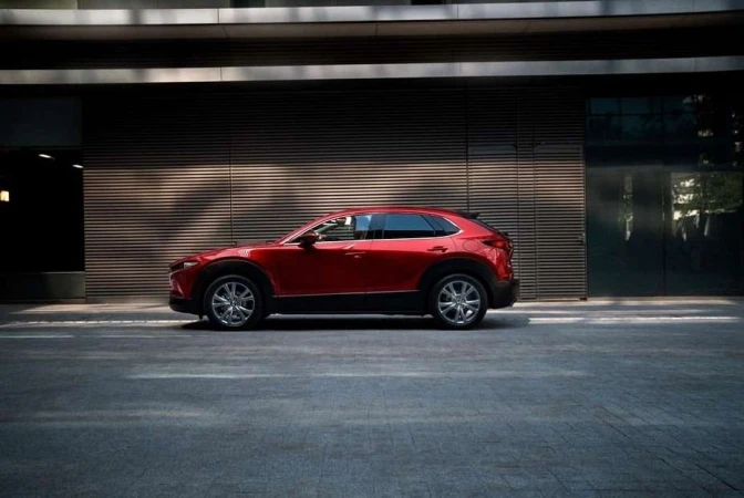 Red Mazda CX-30 parked on asphalt street against a modern building with horizontal lines and glass doors, captured in an eye-level shot.