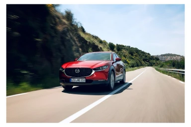 Red compact SUV driving on a winding coastal road with rocky cliffs and vegetation under a clear sky