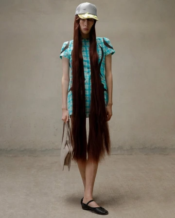 Full-length shot of a woman wearing a blue plaid dress with brown fringe-like hair extensions, a baseball cap, and black shoes in studio.