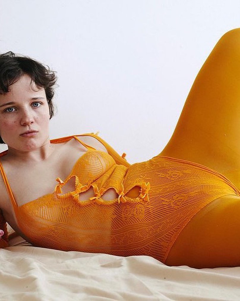 A woman in an orange bodysuit reclines on a white surface, posing in a studio with soft lighting, creating a fashion-forward, relaxed portrait.