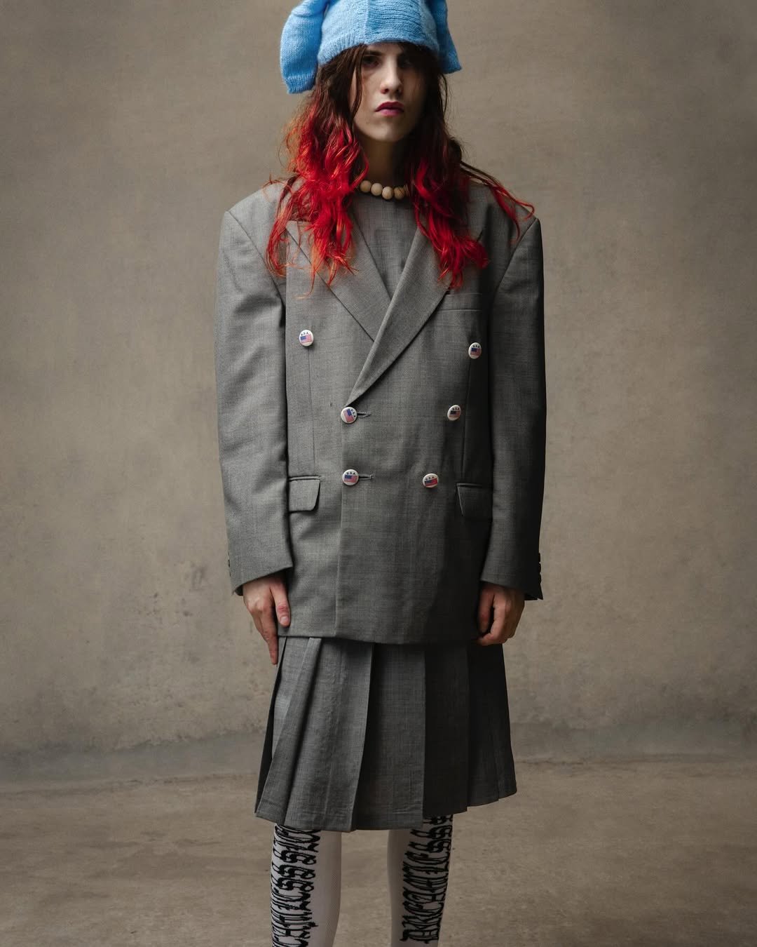 Young woman poses in grey double-breasted suit, pleated skirt, white patterned tights, and a knitted blue hat against a grey backdrop.