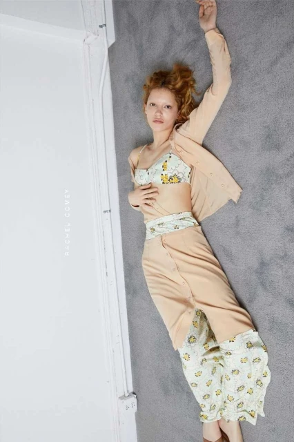 Overhead shot of a woman lying on a gray rug in a light peach two piece floral outfit in a bright studio with white walls.