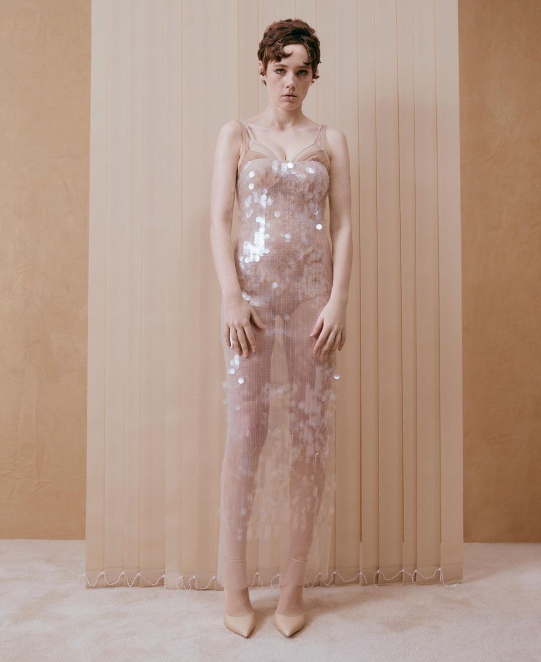 Woman poses in sheer, sequined gown against a neutral backdrop. Full-length shot captures her elegant style in soft lighting.