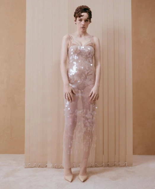 Woman poses in sheer, sequined gown against a neutral backdrop. Full-length shot captures her elegant style in soft lighting.