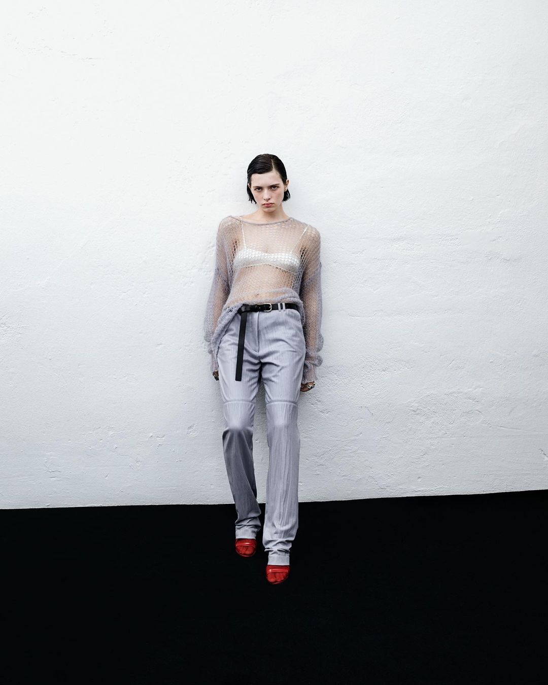 A slender woman with short, dark hair poses in a sheer top and grey trousers, standing against a contrasting grey and black wall indoors.