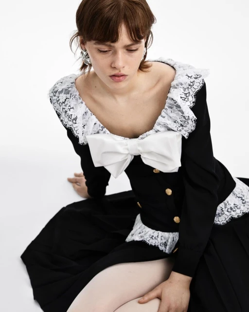 A woman with brown hair sits in a black and white dress featuring lace trim and a large bow on a white seamless background.