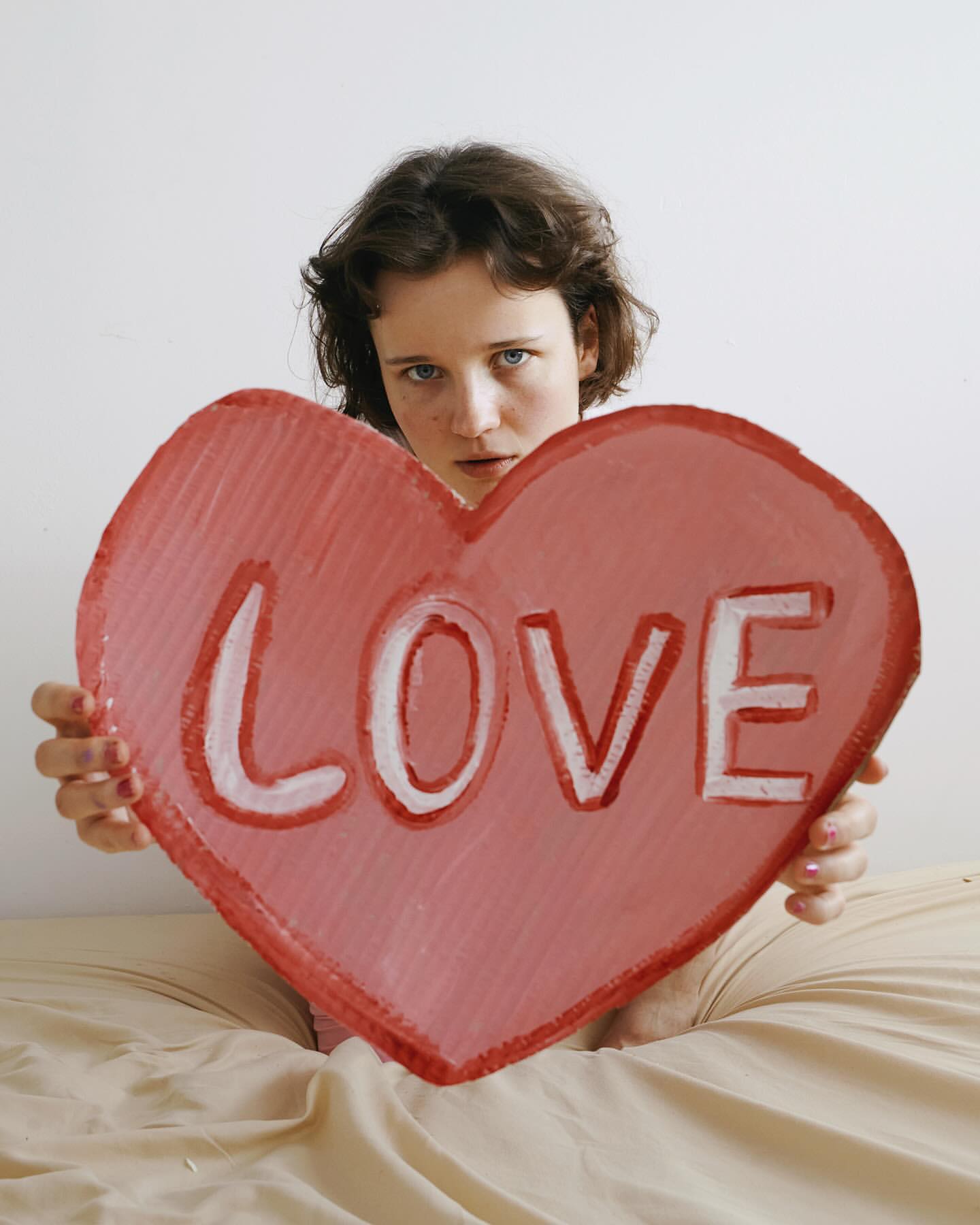 A young woman with dark hair in bed holds a large cardboard heart with the word 'Love' painted on it in a brightly lit, indoor setting.